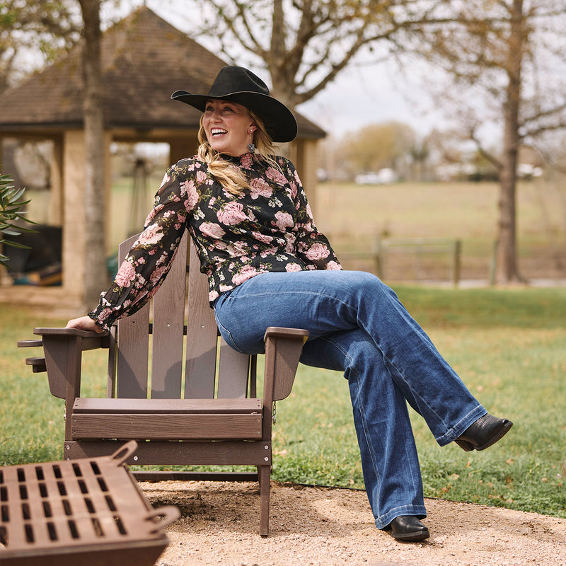 Woman sitting on a wooden chair outdoors wearing a floral shirt, jeans, and black hat, and black Durango cowgirl boots.