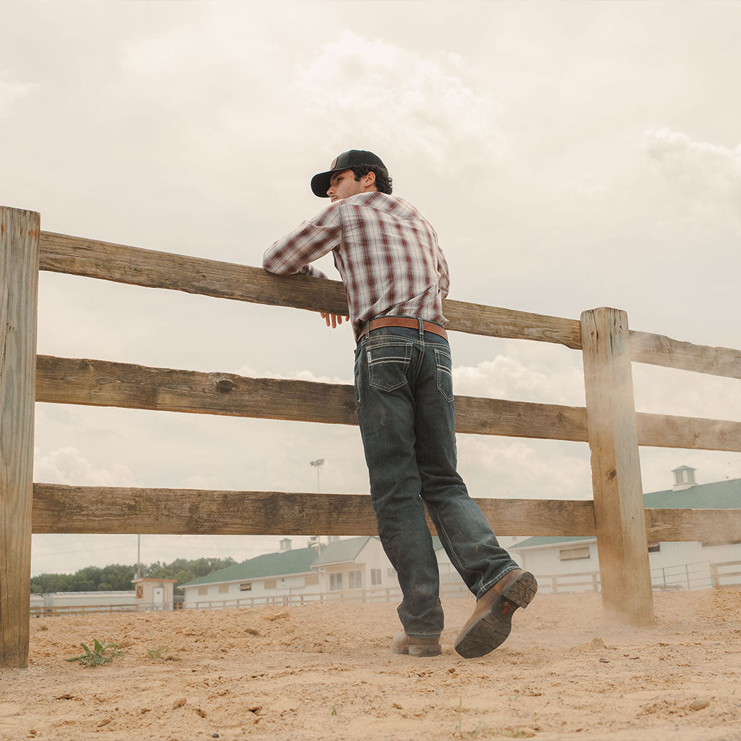 A man in a red and white plaid shirt, dark jeans, and a black cap leans on a wooden fence in a dusty outdoor setting with farm buildings in the background.