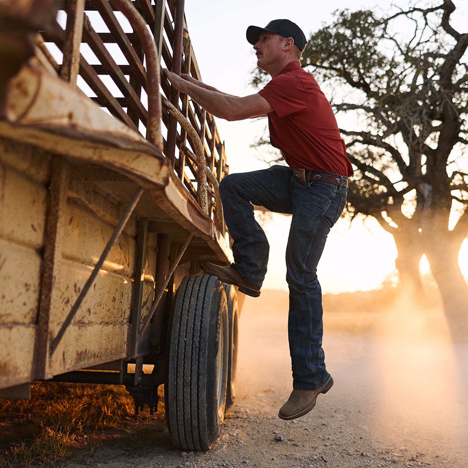 Man climbing into a truck bed on a dirt road with a sunset background wearing durango western boots