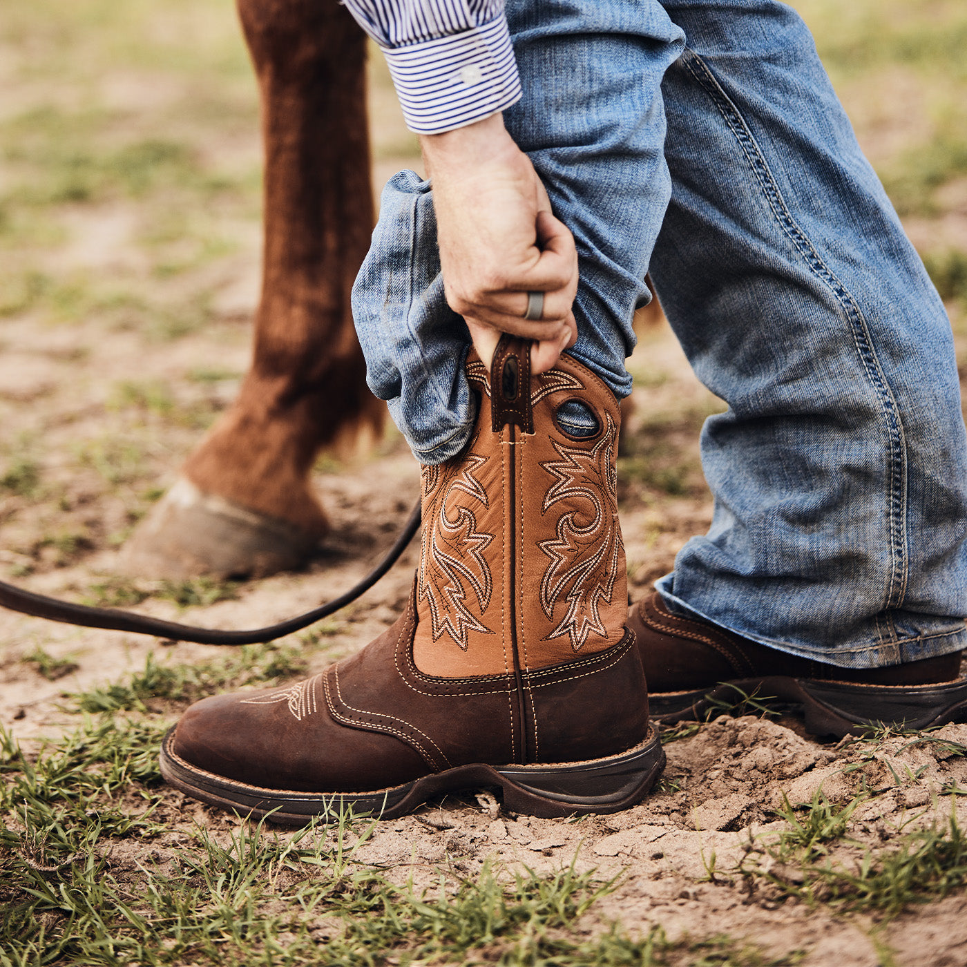 Person wearing brown Durango cowboy boots with intricate stitching, standing on a grassy field with a horse's hoof in the background.