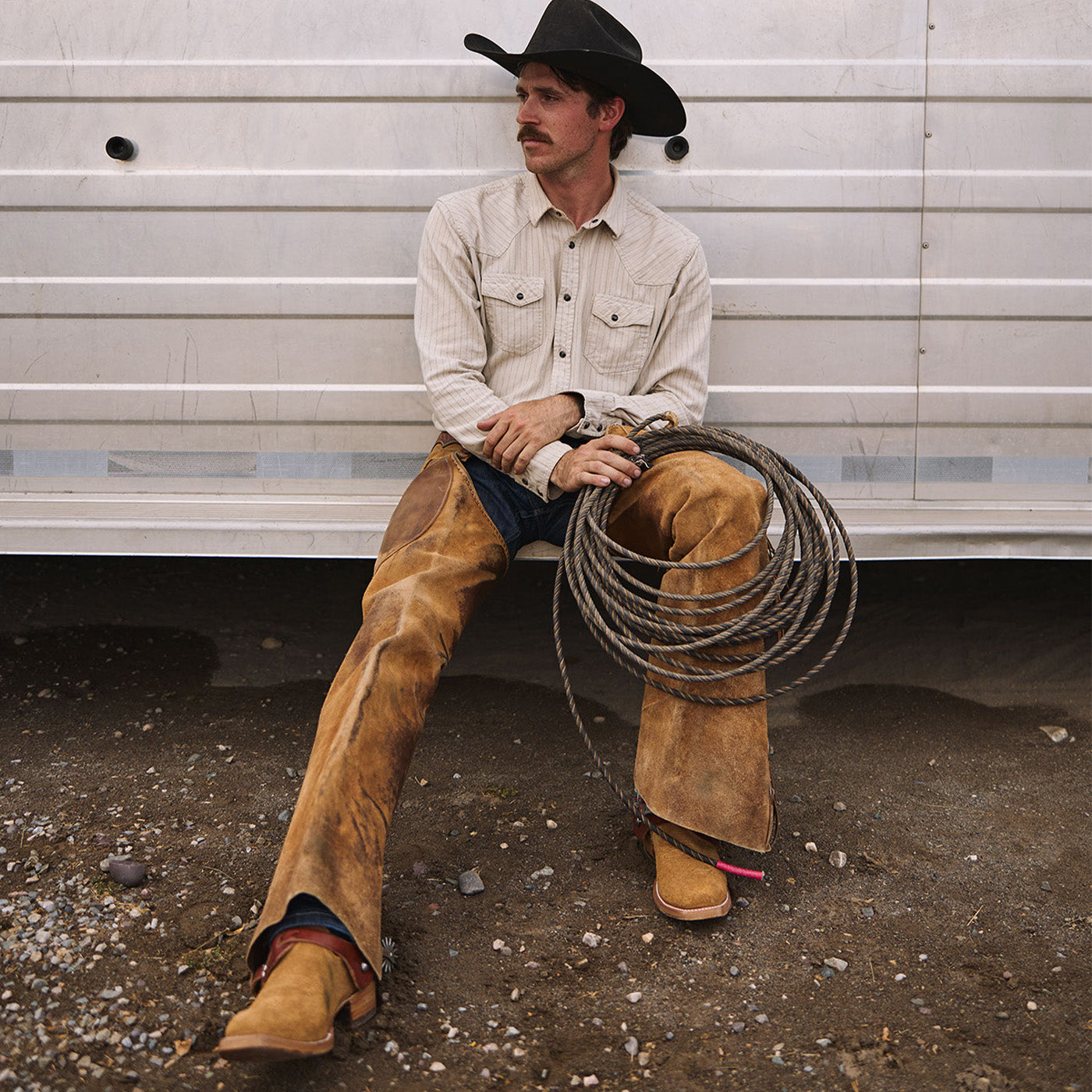Man in cowboy attire sitting on a trailer holding a coiled rope wearing western boots.