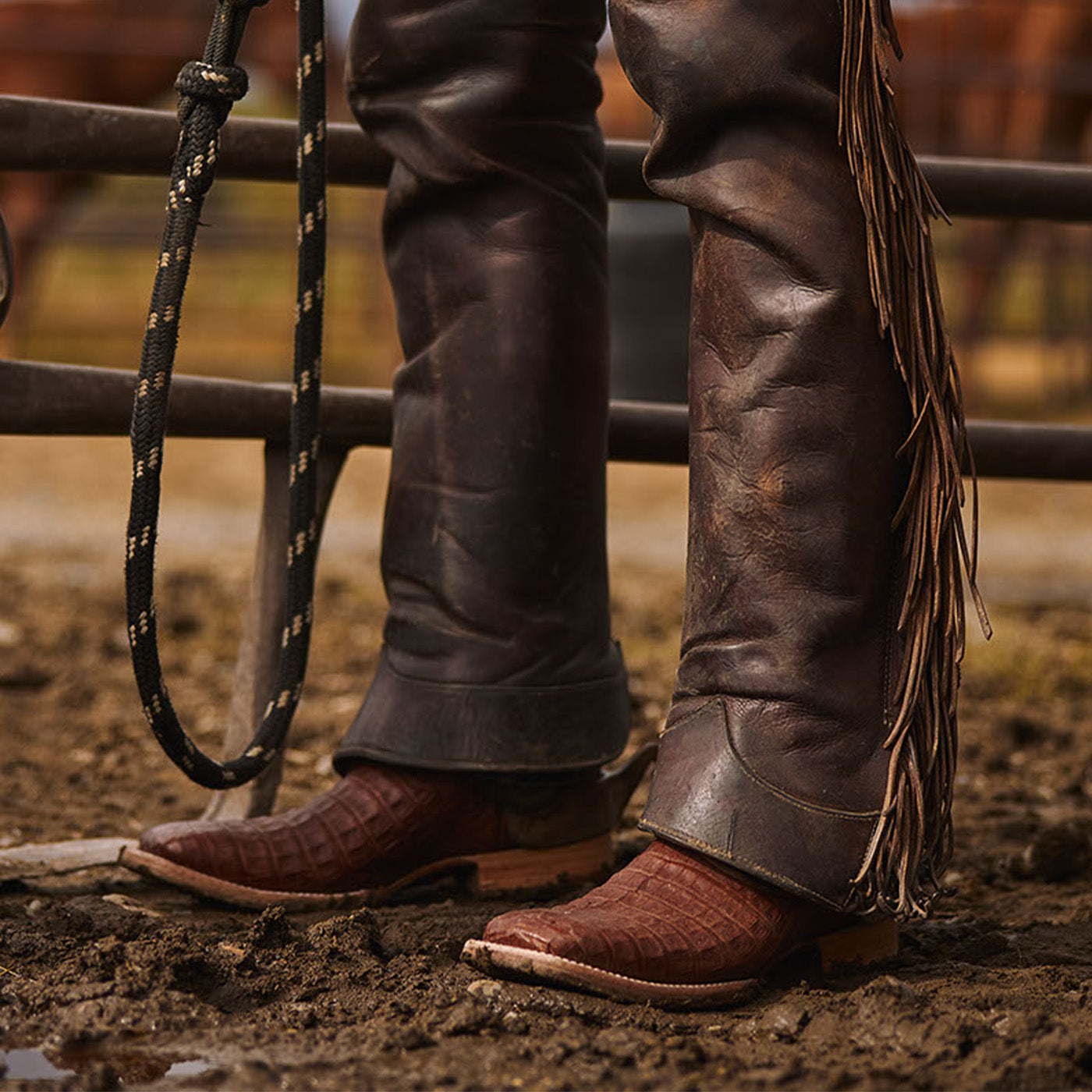 Close-up of a person wearing brown leather cowboy boots with fringes and dark pants, standing on a dirt ground.