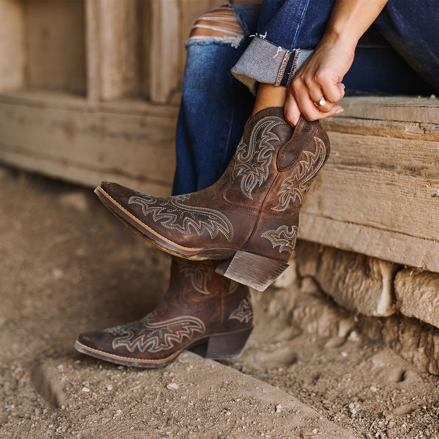 Brown women's short cowboy boots with intricate designs worn by a person sitting on wooden steps.