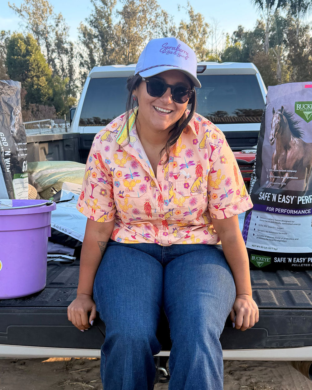 Michelle Serna sitting on a truck bed with a colorful shirt and cap, surrounded by horse-related products.